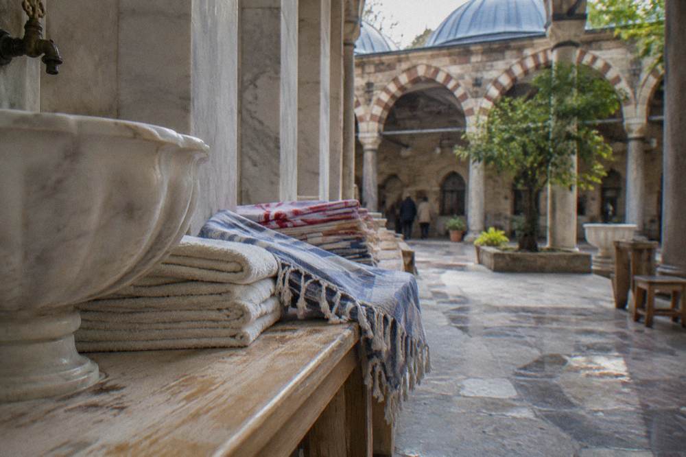Stacked folded towels and woven peştemal cloths on a wooden bench beside a hammam basin