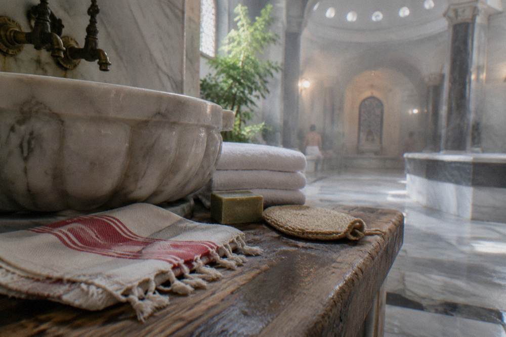 A wooden bench holding a folded peştemal, stacked towels, olive oil soap, and a kese mitt