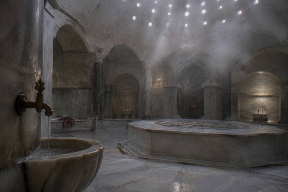 Steam rising in an empty marble hammam chamber with a domed ceiling pierced by light