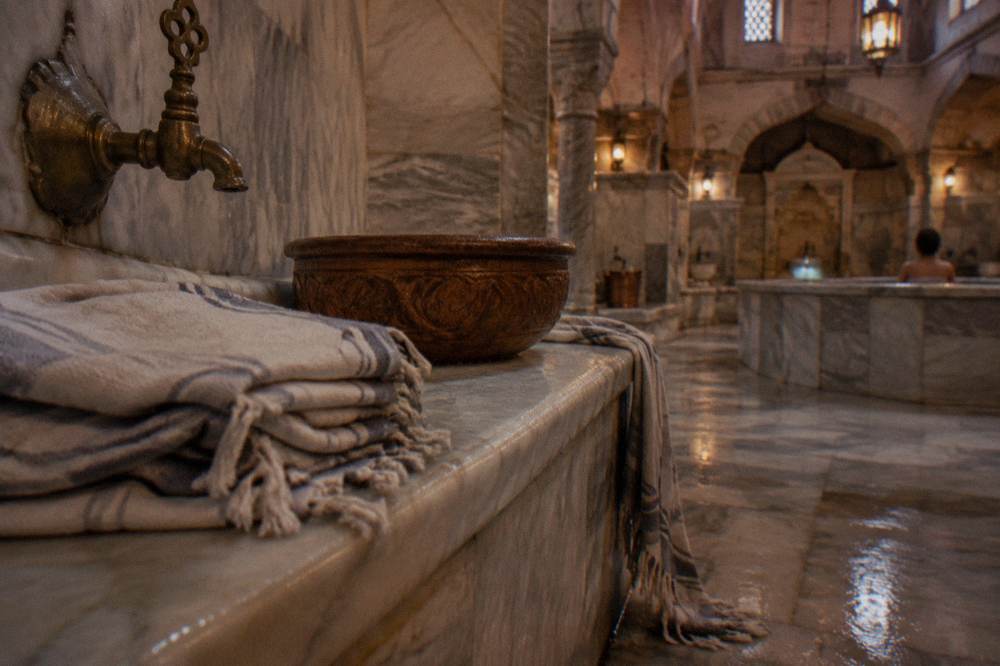 Folded towels and a wooden hammam bowl on a marble bench beside a brass tap