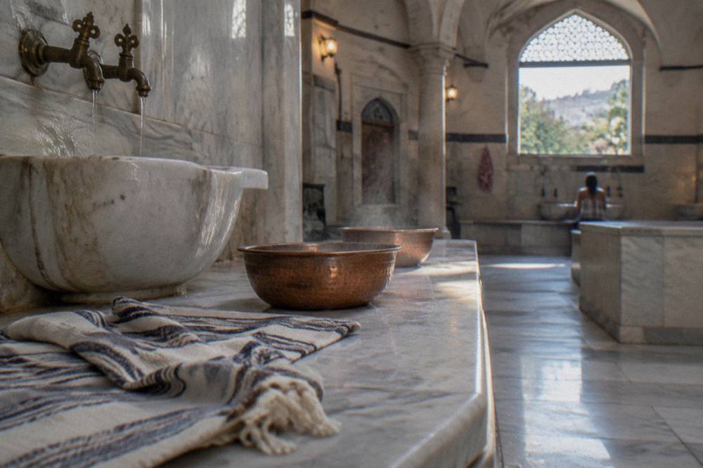Warm marble hamam interior with steam rising, copper bowls, and folded towels on a tiled bench