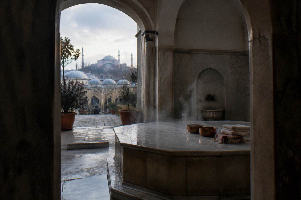 Steam rising in a domed marble hamam with copper bowls and folded towels on a heated stone platform