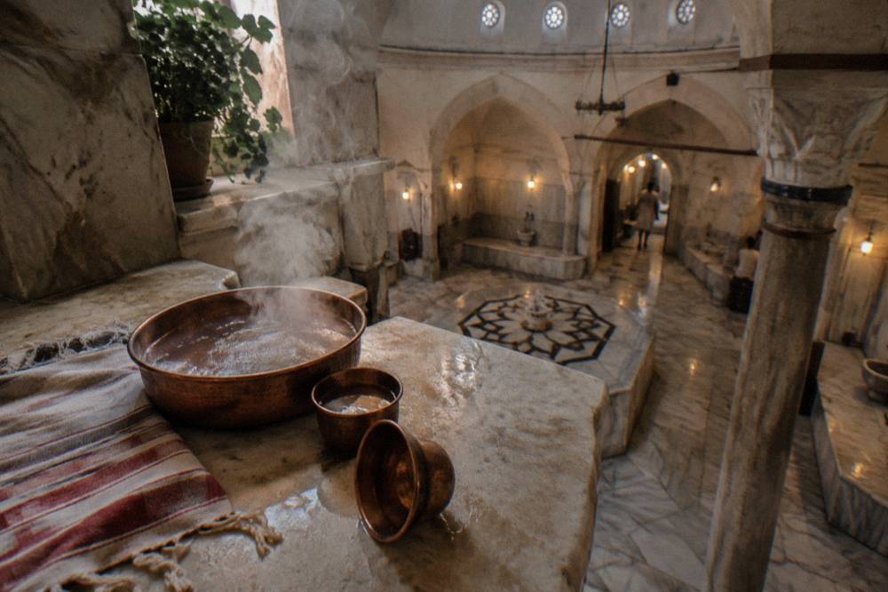 Copper bowls and steam beside a warm marble slab under a domed ceiling in a Turkish hamam.