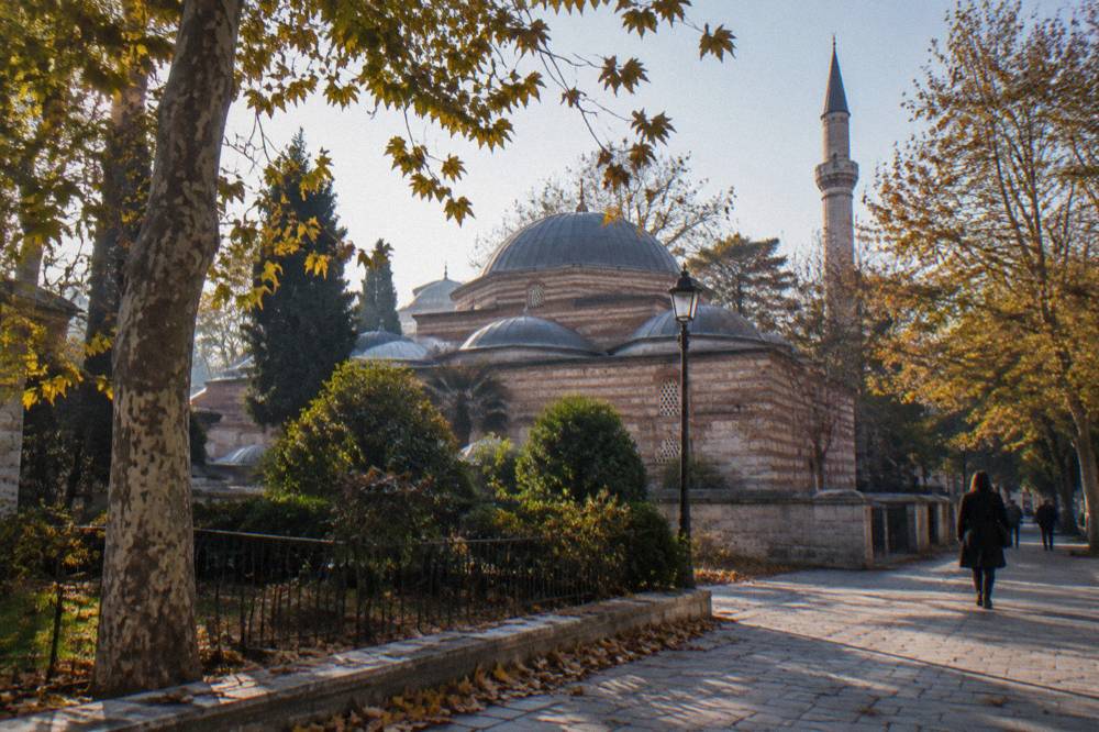 Sunlit plaza in Istanbul with a historic domed bathhouse between tall trees and minarets
