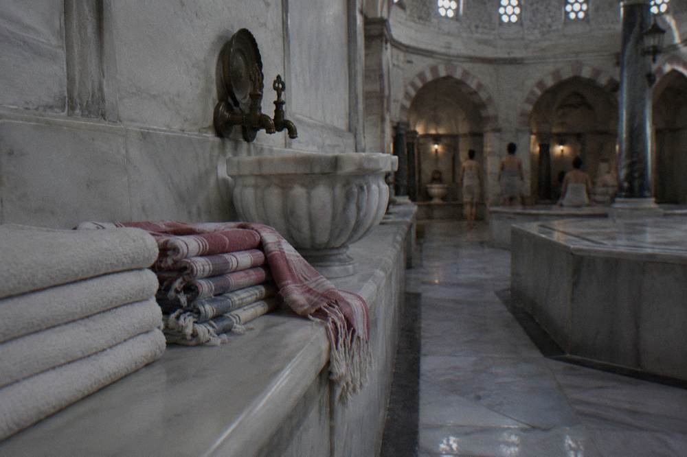 Stacked folded cotton towels and woven peştemal wraps on a wooden bench beside a marble basin