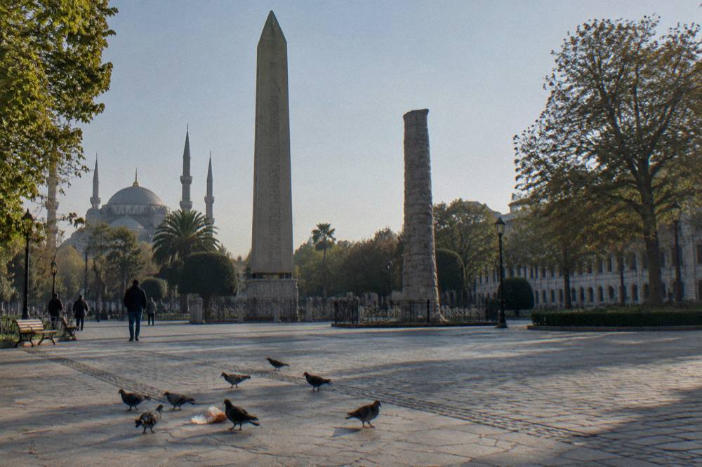Granite obelisk and stone column standing along a wide pedestrian promenade between historic buildings