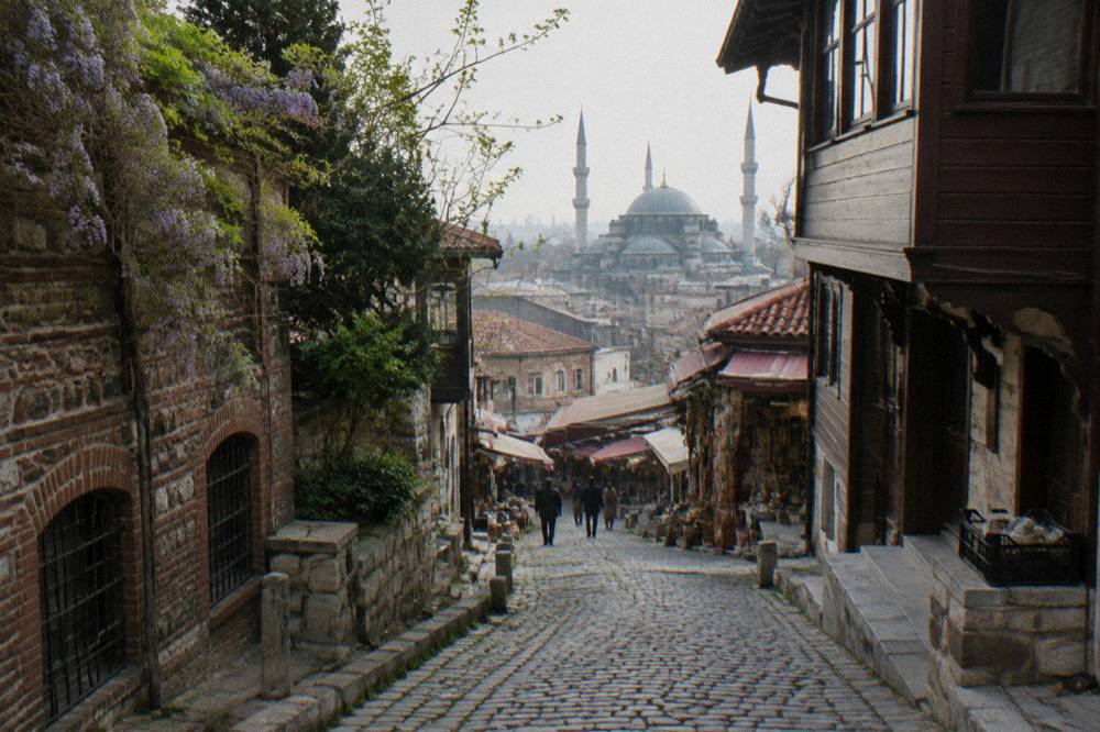 Narrow old street with stone pavements winding between wooden and brick buildings toward a distant mosque dome