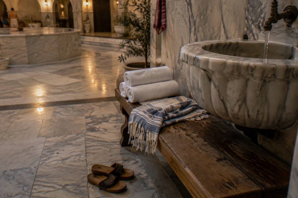 Folded towels, slippers and a woven peştemal on a wooden bench beside a hamam basin
