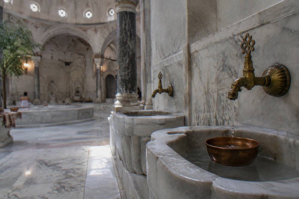 Two adjacent marble basins with brass taps along the wall of a lit domed hall