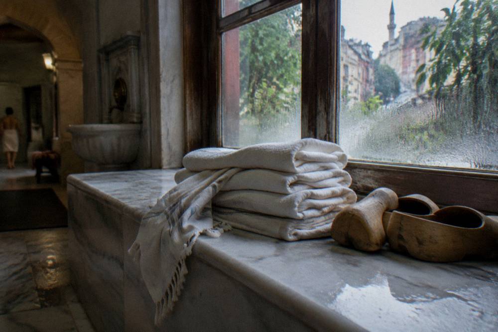 Stacked cotton towels and wooden clogs on a hamam bench beside a steamed window