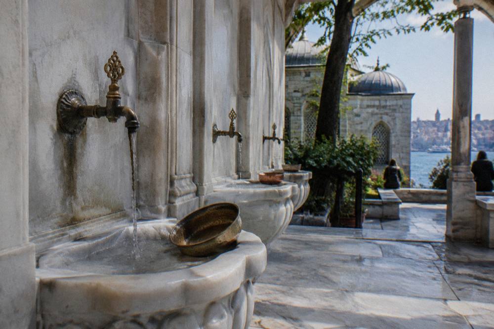 Carved marble basin and copper hamam bowls arranged along the wall of a lit bathing hall